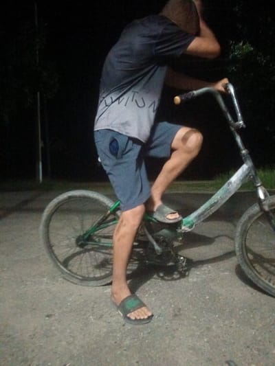 This media file shows a person riding a bicycle at night. The subject is a young male wearing a dark t-shirt with a grey graphic design and dark shorts. He is also wearing dark green flip-flops. The bicycle is old and appears to be a BMX-style bike with a green frame and a silver-colored fork and handlebars. The tires are worn.
The scene is outdoors, with the ground appearing to be a paved or asphalt surface. In the background, there are dark trees and some faint lighting, suggesting it's nighttime. The lighting is dim, creating shadows and a grainy quality to the image. There is no clear indication of weather conditions, but the darkness implies it is not daytime.
The person's posture suggests they are actively riding the bicycle. Their left hand is on the handlebars, and their right arm is bent, with their hand near their head, possibly adjusting their hair or shielding their eyes. One leg is raised, in the motion of pedaling.
The overall mood is casual and perhaps a bit adventurous, given the nighttime setting and the subject's attire. There is no visible text in the image. The location context is Leontyevskoye, Russia.