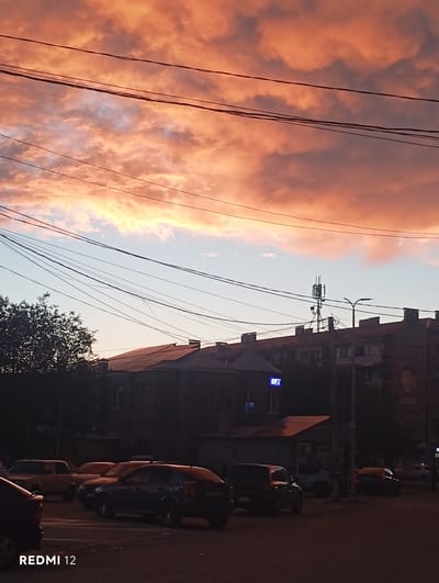 This is a vertical, low-angle shot of a street scene in Vedi, Armenia, captured during sunset. The sky is a dramatic canvas of orange and pink clouds, illuminated by the setting sun. Several power lines crisscross the frame, adding a sense of urban infrastructure against the natural beauty of the sky.

On the right side of the image, buildings line the street. These appear to be multi-story structures with visible windows and architectural details characteristic of urban development. The lower portions of the buildings are partially obscured by shadow. A street lamp is visible atop a pole.

Parked along the side of the street are several cars, their shapes discernible despite the low light conditions. The foreground is dominated by the dark silhouette of a car, and further down the street, more vehicles are parked in a row. A tree with dark foliage stands out against the lighter sky near the buildings.

The overall mood is serene and transitional, reflecting the end of the day. The vibrant colors of the sunset evoke a sense of warmth and natural splendor, juxtaposed with the everyday elements of a streetscape. The presence of the power lines and buildings firmly places the scene in an inhabited area.

The text "REDMI 12" is visible vertically along the right edge of the image, indicating the device used for capture. The time of day is clearly sunset, given the dramatic coloration of the sky. No people are prominently visible in the frame.
