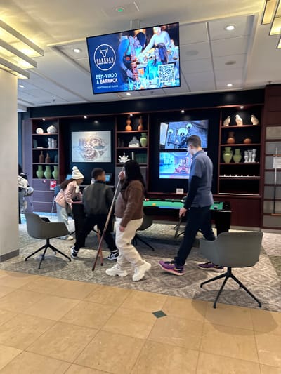 The image depicts an indoor leisure area located within the Claus Hoofddorp complex in Hoofddorp, Netherlands. The central feature is a dark green-felted pool table around which four individuals are present.

On the left, a young girl wearing a pink jacket and white beanie is bent over the pool table, possibly preparing to take a shot. Beside her, a young boy in a dark jacket stands upright, observing the table. To their right, a female wearing a brown sweater and light pants stands, holding a pool cue vertically. Further to the right, a male in a blue short-sleeved shirt over a dark long-sleeved shirt walks from right to left, holding a pool cue.

The background wall is dark, featuring integrated shelving units displaying various decorative ceramic vases and sculptures in light and green tones. A large framed artwork, possibly a blueprint or architectural drawing, is mounted centrally on the wall. Two smaller digital screens are vertically stacked within the shelving on the right, displaying interior renderings.

Above the wall, a large digital screen is suspended from the ceiling, displaying an advertisement for "BARRACA RODIZIO GRILL & BAR" with the text "BEM-VINDO A BARRACA NEXTDOOR AT CLAUS," along with images of food and people.

The flooring in the immediate foreground is light brown tile with dark square accents, transitioning to a patterned grey carpet in the area surrounding the pool table. Several modern grey swivel chairs with black bases are positioned around the table. The space is illuminated by overhead recessed lighting and accent lighting on the shelving units.