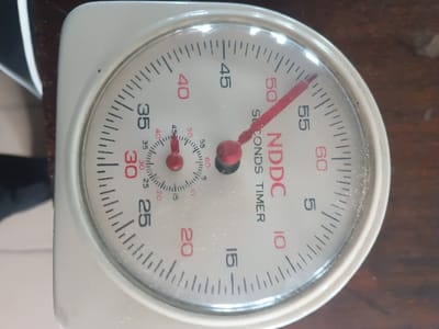 An off-white, analog stopwatch, branded "NDDC SECONDS TIMER" in red lettering, is depicted resting on a dark brown wooden surface. The main dial features red numbers ranging from 5 to 60, indicating seconds, with a prominent red hand currently positioned at approximately 52 seconds. A smaller sub-dial, located at the 12 o'clock position on the main dial, displays black numbers from 5 to 60, likely for minutes, with its smaller red hand indicating approximately 2 minutes. The stopwatch's protective cover is visibly dusted. The background consists of the aforementioned dark wooden surface and an out-of-focus, dark object in the upper left. This scene is located in the city of Warri South, Nigeria.