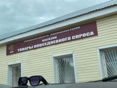 The image shows the facade of a building with a sign in Russian. The sign reads "Самарская область Кошкинское Райпо МАГАЗИН ТОВАРЫ ПОВСЕДНЕВНОГО СПРОСА". This translates to "Samara Region Koshkinsky District Consumer Society STORE OF EVERYDAY GOODS". Below the sign, there are two windows with decorative metal grilles and a logo with the letters "КООП". In the foreground, a pair of dark sunglasses rests on a surface. The building is clad in yellow siding and has a grey metal roof. The sky is overcast. The location is identified as Nadezhdino, Russia.