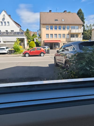 The image shows a street scene in Bad Meinberg, Germany. In the foreground, a dark gray SUV is parked behind a bush. Across the street, a red hatchback is parked next to two yellow trash bins. Further down the street, a white car is parked in front of a building. The building itself is a multi-story structure with a yellow facade and balconies. To the left of the yellow building is a white house with solar panels on the roof. The sky is blue with scattered clouds. The scene appears to be a sunny day.