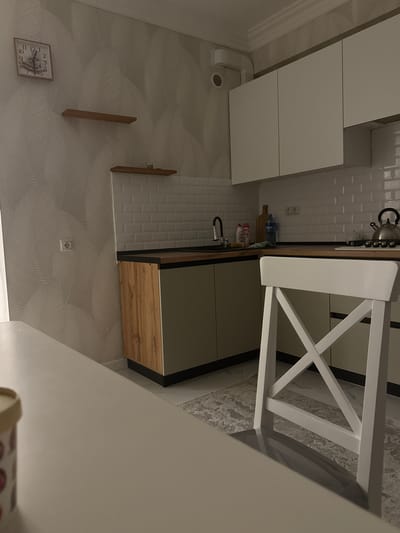 The image depicts a modern kitchen with a light gray, textured wallpaper and white subway tile backsplash. A white chair with a distinctive cross-back design is positioned at a wooden-topped counter. Below the counter, there are light gray cabinet doors and a wooden drawer front, creating a contemporary aesthetic. On the counter, a silver kettle and a few bottles are visible.
To the left of the counter, white cabinets are mounted on the wall, with a ventilation duct visible below them. The wall also features a square clock with Roman numerals. Further to the left, two narrow wooden shelves are mounted on the wallpapered wall. On the right side of the image, the wall has a rough texture and a white electrical outlet is present.
The scene appears to be indoors, likely in a residential kitchen. The lighting suggests it could be daytime or evening, with artificial light sources contributing to the illumination. There are no people visible in the image. The overall impression is one of a clean and stylish, yet lived-in, kitchen space. The specific location is Derbent, Russia, but this is not visually evident from the image itself.