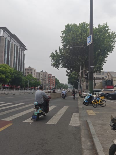 The photograph depicts a street scene in Qinhuai District, Nanjing, China. A man on a blue and white electric scooter is crossing a zebra crossing from left to right. In the background, another individual is riding a scooter further up the street. A woman is walking alongside the scooter rider, pushing a bicycle. Several parked scooters and bicycles line the sidewalk on the right. Behind the scooters, a black car is parked. A tall pole with a sign is visible next to a large tree on the right side of the frame. Buildings of varying heights line the street on both sides. The sky is overcast.