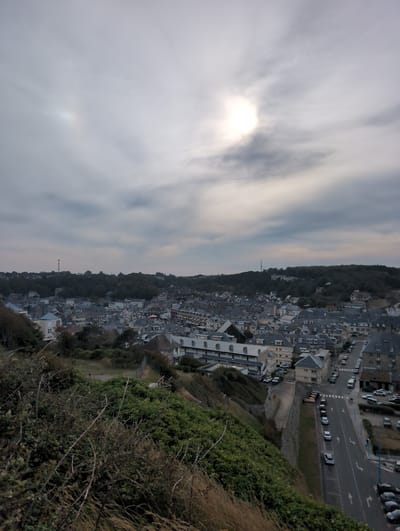 This image depicts a coastal town with a dense residential and commercial area. The buildings are primarily composed of grey slate roofs and stone or light-colored facades, arranged in tightly packed rows. A main road runs through the town, lined with parked cars. In the foreground, there is a steep, overgrown embankment covered in green bushes and dry, thorny branches. The sky is overcast with some areas of brighter light, suggesting the sun is obscured by clouds. A faint solar halo is visible in the upper portion of the sky. The overall impression is of a historic European coastal settlement under a diffused light. The location is identified as Saint-Valery-en-Caux, France.