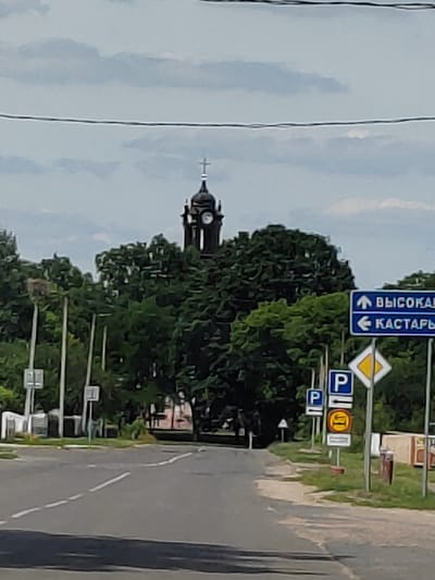 The image captures a street scene in the village of Volchynsky Selsoviet, Belarus. In the background, a dark-colored clock tower with a cross on top rises above a dense canopy of green trees. The clock tower appears to be part of a church or other historic building.
In the foreground, a paved road with white lane markings curves to the right. Along the right side of the road, several traffic signs are visible. A blue sign with a white arrow indicates "ВЫСОКАЯ" (High) to the right, and another blue sign with a white arrow points left towards "КАСТАРЫ" (Kastary). Below these, a diamond-shaped yellow and white sign, likely a warning sign, is present, followed by blue signs with a white letter "P" indicating parking. A circular red and white sign with a symbol of a car might denote a no-entry or specific parking restriction.
On the left side of the road, there are some white poles, possibly for streetlights or utility lines, and some smaller signs. The environment is characterized by a lush green landscape with trees dominating the background and a partially overcast sky. The overall impression is a quiet, semi-rural road leading towards a village center.