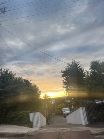 A serene sunset illuminates a cloudy sky above a residential street. Silhouetted trees and power lines frame the left side, while houses and parked vehicles line the right. Golden light pierces through the clouds, creating a striking contrast against the darker foliage and buildings. The image captures the tranquil ambiance of twilight in a suburban setting.