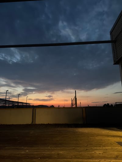 The image depicts a dusky sky over Singen (Hohentwiel), Germany, with a dramatic cloud formation obscuring most of the sunset. The foreground consists of a wooden deck, partially illuminated by a warm light source coming from the right. Beyond the deck, a low, light-colored wall with dark vertical supports separates it from a darker area, possibly a courtyard or a lower level. In the mid-ground, a fenced structure, possibly a sports court with a volleyball net, is visible. Streetlights stand in a row to the left, and distant hills are silhouetted against the horizon. A modern building with horizontal siding is visible on the far right edge of the frame. The scene appears to be an outdoor space, possibly a terrace or viewing area, overlooking a public or recreational facility. There are no people or animals visible in the image.