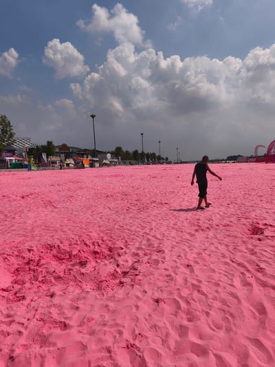 A man walks across a vast expanse of bright pink sand under a bright blue sky with scattered white clouds. He is wearing a dark sleeveless shirt and shorts, and appears to be walking away from the camera towards the right side of the frame. The pink sand is textured with footprints and small mounds. In the background, a row of buildings and structures are visible, including what appear to be vendor stalls and some tents or event structures. Trees and lampposts are also scattered in the distance. The overall impression is that of an artificial beach or an outdoor event space in an urban setting. This location is in Nanguan District, China.
