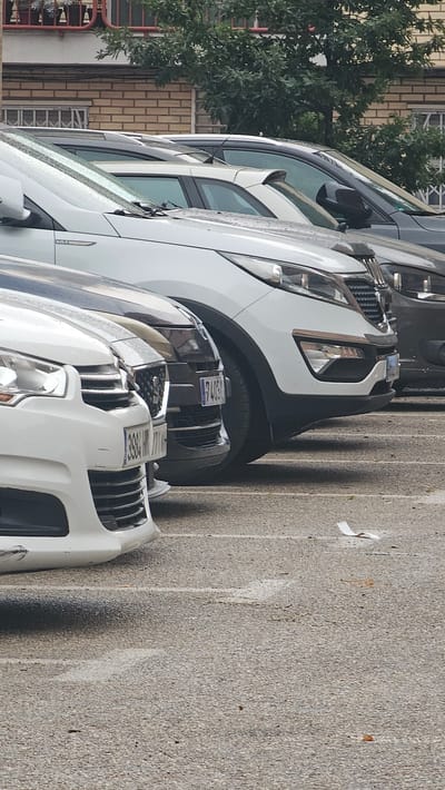The image shows a row of parked cars on a paved surface, likely a street or parking lot, in Zaragoza, Spain. The cars are oriented vertically in the image, suggesting the photo was taken with the camera held sideways or the image was rotated. The closest car in the foreground is white, with a black grille and headlights visible. Further back, a white SUV-like vehicle is parked, with its front bumper and grille visible. Behind that, another white vehicle, possibly an SUV, is parked. Further still, a dark-colored vehicle is visible, with its side mirror and part of its body showing. The background on the left shows a brick building and lush green trees. The ground is a textured gray asphalt. There are faint white lines on the pavement, suggesting parking spaces or lane markings. Some small pieces of white paper are scattered on the ground.

The activity appears to be simply that of parked cars. No people are visible in the frame. The lighting suggests it is daytime, with shadows cast by the cars. The presence of water droplets on the car windshields indicates recent rain or a wet environment. The license plates of the cars are partially visible and show alphanumeric characters, but are not clearly legible for full identification. The overall scene is mundane and typical of an urban or suburban parking area.