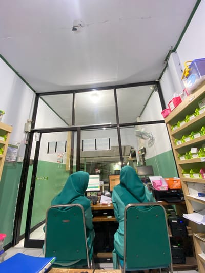 This media file shows an interior view of what appears to be a pharmacy or a medical clinic, likely located in Margoyoso, Indonesia. The scene is shot from a slightly elevated, wide-angle perspective, with the camera oriented sideways.
Two individuals, both wearing teal-colored hooded smocks or uniforms, are seated at desks, facing away from the camera. They are positioned behind a glass partition, suggesting they are working in a customer-facing area or an administrative section. One person is clearly visible at a computer monitor with a keyboard. The other is partially obscured by the first person and their chair.
The setting includes shelves stocked with various items, possibly medical supplies or documents, with some items in red and orange baskets. A ceiling fan is visible above the desks, and there are computer monitors and printers on the desks. The walls are painted white and green.
Visible text includes signs with Indonesian words. On the green door, a sign reads "GESER" with an arrow, meaning "slide" in Indonesian, indicating how to open the door. Another sign on the wall above the desks shows a row of text, some of which is obscured.
The lighting suggests it is daytime or artificial lighting is in use, creating a bright interior. The overall impression is one of a functional, busy workplace.