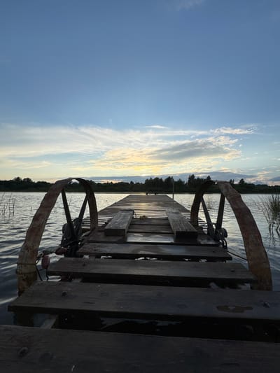 The image shows a wooden pier extending over a body of water. The pier is weathered and appears to be made of dark planks. On either side of the pier, towards the foreground, are two rusted, curved metal structures, possibly part of a winch or mooring system. The water is calm with gentle ripples. In the background, a line of trees borders the water under a sky with scattered clouds, tinged with the colors of sunset. The overall scene suggests a natural, tranquil environment, likely a lake or slow-moving river, possibly in or near Sitno, Russia, given the vegetation and water. There are no people or animals visible in the image.
