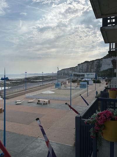 The image depicts a seaside promenade in Saint-Valery-en-Caux, France, featuring a beach, a casino building, and apartment complexes. The sky is overcast with scattered clouds, and the sea is visible in the background. On the promenade, there are lounge chairs, picnic tables, and a sandy area with a volleyball net. A blue beach hut and a white umbrella are also present.
To the right, a portion of a dark blue balcony railing is visible, adorned with a yellow planter containing pink flowers. The casino building is situated in the middle ground, its name clearly displayed in large white letters. Behind the casino, steep white cliffs rise up, characteristic of the region. Several cars are parked in front of the casino, and a few people are walking on the promenade and the beach. The overall atmosphere is calm and appears to be during daylight hours.