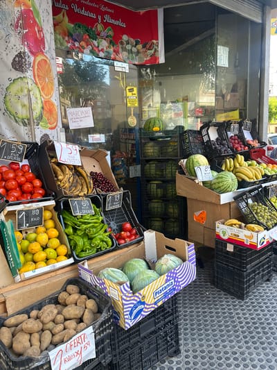 This is a vertical, eye-level, medium shot of a fruit and vegetable stand outside a store in Castelló De La Plana, Spain. The stand is overflowing with produce, including watermelons, bananas, grapes, plums, tomatoes, peppers, lemons, potatoes, and cabbage. Some of the produce is displayed in black plastic crates, while others are in cardboard boxes. Signs with prices and the word "OFERTA" (offer) are visible throughout the stand.
In the background, the store's interior is visible through a large glass window. Store shelves are lined with packaged goods, and some of the interior appears to be a refrigerator case. A red banner on the left side of the store entrance features an illustration of various fruits and vegetables and text in Spanish that reads "Productos Latinos Frutas Verduras y Alimentos." Another sign on the store window reads "ATENCION ZONA PROTEGIDA POR CAMARAS DE VIGILANCIA LAS 24 HORAS," indicating a surveillance zone.
A sign handwritten on white paper is taped to the store window and reads "MARCIANO De lucmay Guenabana/Fresa a 150 y mango." This likely refers to specific fruit varieties and their prices. The scene appears to be outdoors during the daytime, with bright sunlight casting shadows. The ground is paved with gray circular tiles. The overall impression is of a bustling market scene, with a focus on fresh produce and local offerings.