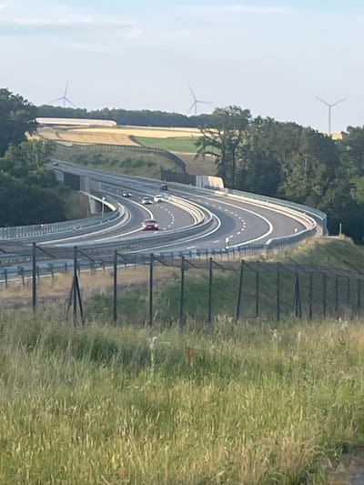 The image depicts an elevated highway curving through a landscape with wind turbines in the distance. The highway has multiple lanes with cars driving on it. The surrounding area is covered in trees and fields. In the foreground, a grassy field with a fence is visible. The sky is partly cloudy. The specific location is the A7 highway near Momberg, Germany, in the vicinity of the Amberg and Schnaittenberg wind farms.