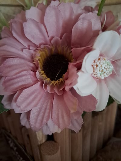 This is a close-up, eye-level, indoor photograph of artificial flowers. The dominant flower is a large, light pink daisy-like bloom with a dark brown center and yellow accents. To its right is a smaller, white flower with a textured, bumpy center. The flowers appear to be in a wooden planter, indicated by vertical wooden slats visible at the bottom of the frame. The background is blurred, suggesting a shallow depth of field, and includes a textured surface, possibly a wall. There are no people or actions depicted. The location in Gavar, Armenia, is not evident from the image itself.