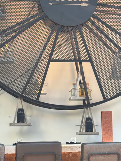 Here's a detailed description of the image:
The image features a close-up view of an interior scene, predominantly showcasing a decorative metal structure. This structure is circular with a radiating pattern created by metal rods intersecting within a perforated sheet metal frame. The design gives off a modern and industrial aesthetic.
Within the circular metal frame, a set of shelves are visible, displaying liquor bottles. The bottles are of various sizes and colors, adding visual interest to the composition.
The setting appears to be a retail establishment, possibly a liquor store or a bar, as evidenced by the display of alcohol and the contemporary design elements. To the right of the frame, there are signs and what appear to be display cases or shelves with merchandise, seemingly related to the liquor or bar setting.
Based on the context of the provided location data (Huntington Beach, United States of America), it can be inferred that this image was taken inside a business within this city, perhaps a store selling liquor or a similar establishment with a focus on beverage retail.