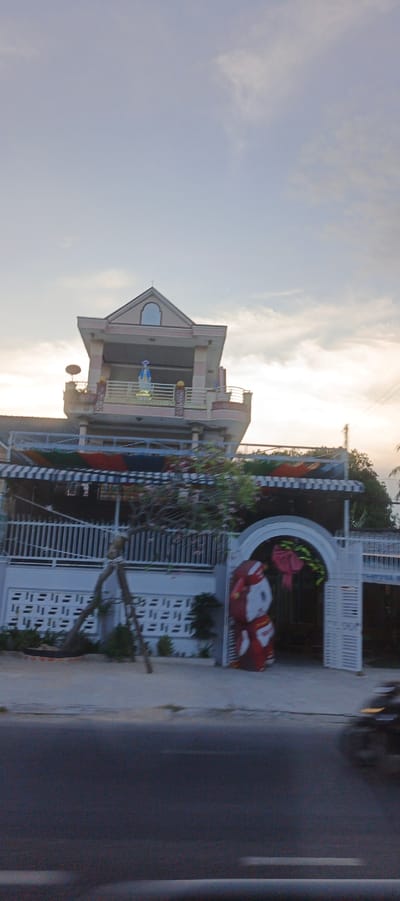 The image shows a multi-story house with a statue of the Virgin Mary on the top balcony, located in Cam Ranh Ward, Vietnam. The house has a peaked roof with a small window. The balconies are decorated with railings. In front of the house, there is a white fence and an arched gateway. A large figure dressed in red and white, possibly a Christmas decoration, stands by the gateway. There is a tree with pink flowers in front of the fence. The scene is set against a cloudy sky with the sun setting or rising. A blurred image of a vehicle is visible on the road in the foreground.