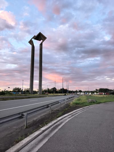 The image depicts an outdoor scene at dusk in the city of Saint-Aybert, France. In the foreground, a paved road curves to the right, bordered by a concrete curb and a metal guardrail. Beyond the road, a grassy area is visible, with some scattered rocks. In the midground, two tall, slender concrete structures rise vertically, topped with a dark, abstract geometric shape. These structures are positioned on the left side of the image. Further back, a multi-lane highway stretches into the distance, with a few vehicles, including trucks, visible. The background is dominated by a dramatic sky filled with clouds tinged with pink and purple hues, indicative of sunset. Trees and some streetlights are sparsely distributed throughout the scene, suggesting an area on the outskirts of a town or a roadside location. The overall impression is one of an urban or semi-urban landscape at twilight.