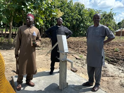 The image depicts three men standing outdoors in Afogo, Nigeria, near a hand pump. The man on the left is wearing a brown tunic and pants, a red cap, and brown loafers. He is holding a small green object and a tool that resembles a hoe. The man in the center, dressed in a black clerical shirt with a white collar, glasses, and black pants, stands with his hands on his hips. The man on the right is wearing a grey tunic and pants and brown sandals, also with his hands on his hips. They are positioned on a concrete pad surrounding the grey metal hand pump, which is situated in a dirt field with some vegetation and scattered small buildings in the background. The weather appears to be clear with a partly cloudy sky.