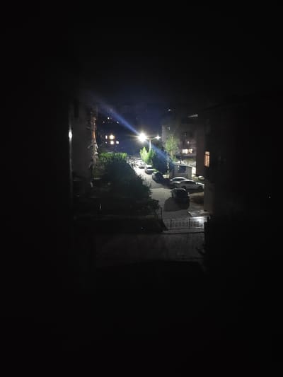 The image depicts a nighttime scene on a street in Tursunzoda, Tajikistan. The view is from a window, looking out onto a street lined with buildings and trees. A streetlamp illuminates the scene with a bright, bluish light, casting long shadows. Several parked cars are visible on the street. The buildings have illuminated windows, suggesting occupancy. The overall impression is that of a quiet residential street at night.