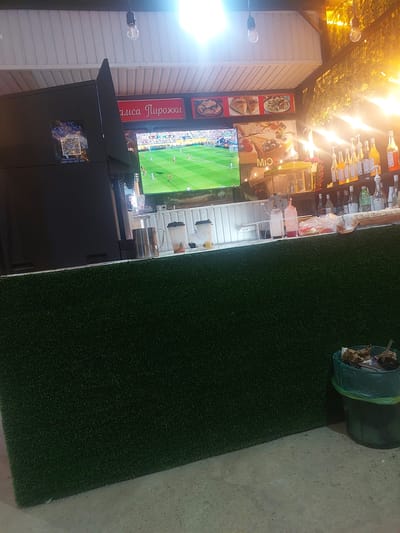 The image shows a food stall or cafe with a television screen displaying a soccer match. The stall has a green, artificial grass-covered counter. Behind the counter, there are shelves with bottles of syrups and alcohol, as well as a coffee machine. A digital display and a QR code are visible on the left side of the stall. In the foreground, there is a trash can filled with debris. The scene appears to be indoors or under a covered outdoor area, with artificial lighting. The location is identified as Ması, Kyrgyzstan.