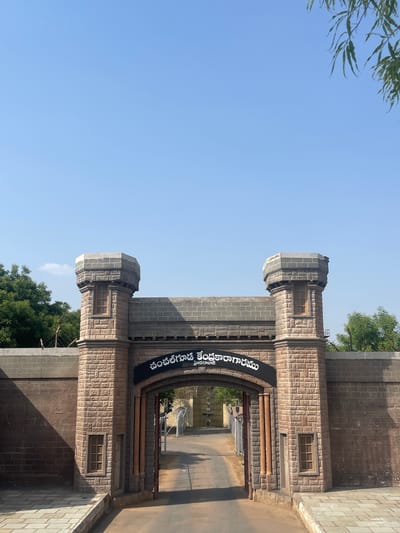 The image shows the entrance to the Central Jail in Anajpur, India. The entrance is a stone archway with two towers on either side. A sign in a Telugu script is located above the archway. The view through the archway reveals a path leading into the jail complex. The sky is blue, and there are trees visible in the background. The scene appears to be taken during daylight hours.