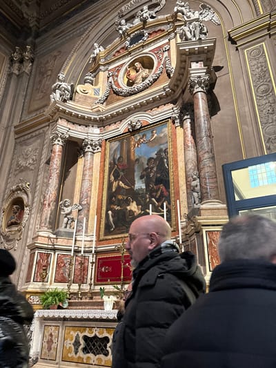 The image captures the interior of the Church of Santa Maria in Vado, Ferrara, Italy, focusing on a prominent Baroque altar structure. The central element is a large altarpiece featuring an oil painting, "The Martyrdom of Saint Maurelius" by Guercino, depicting a multi-figure scene of martyrdom, with soldiers and a bound figure in the foreground and cherubs in the sky above. This painting is set within an elaborate architectural frame composed of pairs of fluted, reddish-brown marble columns, white marble sections, and gilded decorative elements. Above the painting, the altarpiece features a curved pediment adorned with numerous white marble or stucco sculptures of angels and cherubs, with a bust of a saint in a circular medallion at its apex.
Below the painting, the altar table is covered with a white lace cloth. On the altar, multiple tall white candles are positioned in ornate brass candlesticks, accompanied by small potted plants and a white decorative vase. The altar frontal is paneled with ornate marble inlays. To the left of the main altar, a wall niche displays a relief bust labeled "S. PETRUS." The surrounding walls feature stucco reliefs and gold trim.
In the foreground, slightly right of center, a bald male with glasses, a dark puffer jacket, and a patterned scarf is depicted in profile, facing towards the right side of the altar. To his immediate left, a person wearing a dark, possibly glossy, jacket is partially visible. On the far right, another individual with gray hair, wearing a dark jacket, is partially visible from behind. A modern-looking, dark-framed glass structure is present on the far right edge of the frame.