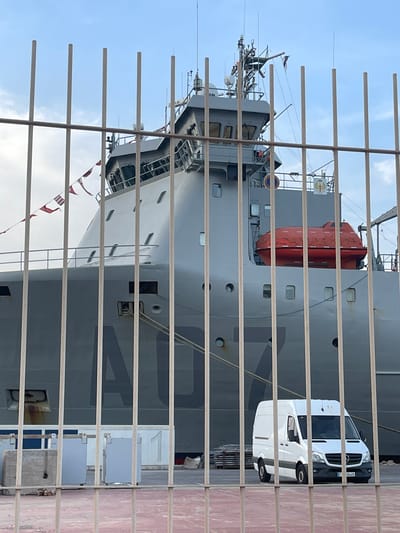 A ship is docked in Cartagena, Spain, with a white van parked nearby. The ship is large and gray, with a red life raft mounted on its side. The van is a Mercedes-Benz Sprinter with a license plate that reads "E 8448." The image is taken from behind a metal fence, which partially obscures the view of the ship and the van. In the background, the sky is cloudy. The ship has the markings "A07" on its side.