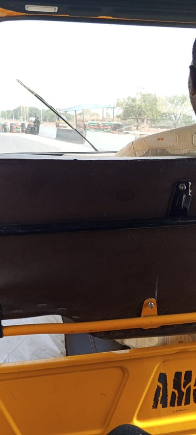 The image shows a view from inside a yellow auto rickshaw, commonly used as transportation in Nigeria. Outside the vehicle, the scene features a military or security checkpoint near Uke, Nigeria. This is indicated by the presence of uniformed personnel, tires forming barriers, sandbags, and a guard post structure. A road roller vehicle is also visible. Inside the rickshaw, the view is partially obstructed by the vehicle's brown interior paneling and structural elements.
