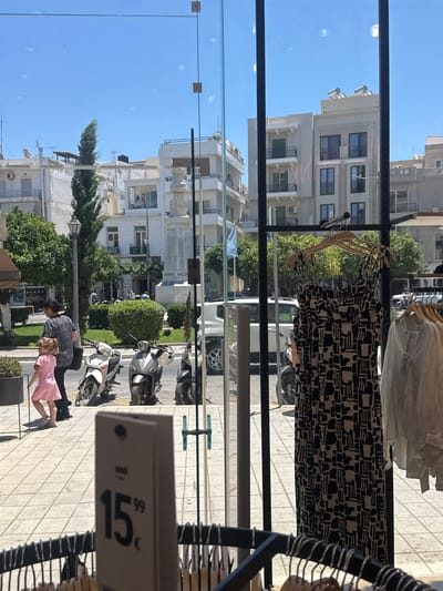 This media file captures a street scene through a storefront window, likely in Agios Nikolaos, Greece, on a bright, sunny day. The primary subjects are two people, a woman and a young girl, standing on a tiled sidewalk outside the store. The woman, wearing a patterned dark top, is bent slightly, seemingly interacting with the child. The young girl, in a pink dress, is also bent over, facing away from the camera.

Inside the store, visible through the glass, are several garments hanging on display racks. A black and white patterned dress is prominently featured on a hanger. In the background, the street is lined with buildings, typical of a Mediterranean urban environment, with balconies and windows. A white van and several scooters are parked along the roadside, suggesting a relatively busy urban area. Green trees and bushes add to the scene's liveliness.

A notable detail is a price tag visible in the foreground, showing "15.99€". The sunlight indicates it is daytime, and the clear blue sky suggests fair weather. The overall atmosphere appears casual and ordinary, a snapshot of daily life on a sunny day in a Greek town.