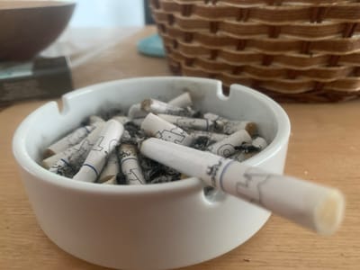 The image shows a white ceramic ashtray filled with many cigarette butts. The ashtray is positioned on a wooden table. In the background, a woven basket is visible, as well as a blurred brown bowl. The lighting suggests it could be daytime, with natural light illuminating the scene. The overall impression is one of a used ashtray, implying someone has been smoking recently. There is no visible text. The scene is set in Tsodoreti, Georgia.