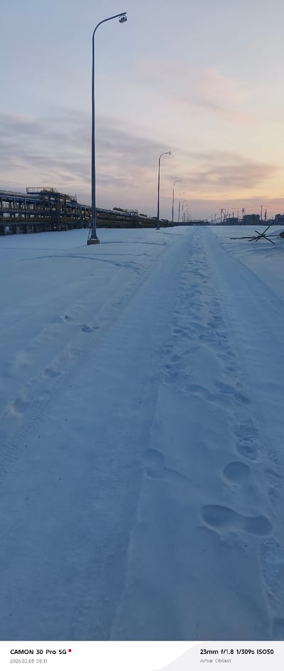 A snowy landscape features a path covered in footprints and tire tracks leading into the distance. Industrial structures stand to the left, under a sky displaying hues of dawn. Streetlights line the road, diminishing in size as they recede into the horizon. The camera details are visible, including a "CAMON 30 Pro 5G" stamp, a date of "2026.02.03 08:11", and technical information like "23mm f/1.8 1/309s ISO50" taken in Amur Oblast.