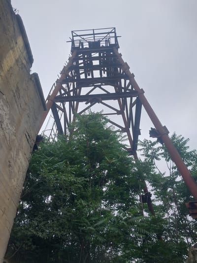 The image depicts an old industrial structure, likely a mining headframe, in the city of Kapan, Armenia. A concrete wall is visible on the left side of the frame, showing signs of age and wear, with exposed rebar. Adjacent to the wall, a large, skeletal metal tower dominates the upper portion of the image. This tower is constructed from rusted metal beams and supports, forming a triangular lattice structure that tapers towards the top. Several pipes run vertically along the tower, also showing signs of rust and some damage, particularly a broken section on the rightmost pipe. Dense green foliage, likely trees and bushes, grows in front of and around the base of the metal tower, partially obscuring it. The sky is overcast and a pale gray. There are no people or animals visible in the scene. The overall impression is one of decay and nature reclaiming an abandoned industrial site.