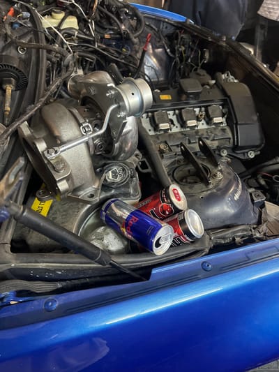 The image displays the open engine bay of a blue passenger vehicle, likely situated in a garage or workshop environment. The specific location within the city of Bersin, Bulgaria, cannot be determined from the visual evidence.
A metallic turbocharger unit, appearing new or recently installed, is prominently positioned on the left side of the engine compartment. It features a visible wastegate actuator and associated mounting hardware. The vehicle's engine block and a black valve cover are partially visible to the right, displaying multiple ignition coils and accompanying wiring.
On the right side of the engine bay, resting on a structural component, are three opened or partially consumed energy drink cans: one blue Red Bull can and two red cans labeled "Black Wings Classic" with a devil-head logo. A yellow adhesive label with the partial text "ALL STO" is visible on a dark surface to the left of the turbocharger. Various hoses, electrical conduits, and other mechanical components are dispersed throughout the engine bay, indicating an active state of disassembly, assembly, or repair. A black shock absorber or strut component is visible extending into the lower left of the frame. No individuals are present in the image.