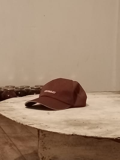 A maroon baseball cap with white embroidered text "BOVERAT" rests on a rustic wooden surface in the foreground. Behind the cap, a row of glass jars with dark contents and metal lids are visible. The background is a plain, textured wall. The image appears to be taken indoors, possibly in a home or storage area, with warm, dim lighting. The scene is static and focuses on the cap and jars. There is no indication of the city of Көкпекті, Kazakhstan within the image itself.