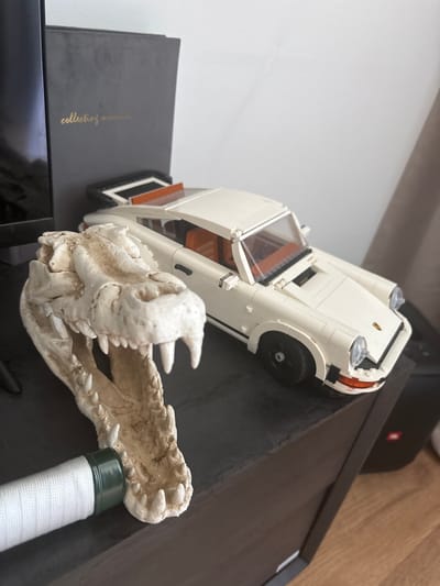 This media file shows a close-up, angled shot of a white Lego car, possibly a classic sports car model, positioned on a dark surface. The car is white with clear windows and orange interior details. A decorative replica of a dragon or monster skull with sharp teeth is placed in front of the car, appearing to loom over it. The skull is pale, textured, and shows intricate detail in its bone structure. To the right of the skull, a white object with a green cap is partially visible, wrapped in white cord, resembling a handle or grip.
The background includes a light gray vertical surface on the left and a wooden surface and a black electronic device with a red logo on the right. The overall setting appears to be indoors, possibly on a shelf or desk, given the arrangement of objects and the background elements. The lighting suggests it is daytime, but no specific time of day is discernible. There is text visible on a dark grey book or box underneath the car, which appears to read "collecting..." in gold lettering, though it is partially obscured. The scene is static, with no activity taking place other than the arrangement of the displayed objects. The location context provided is Gebersdorf, Austria.