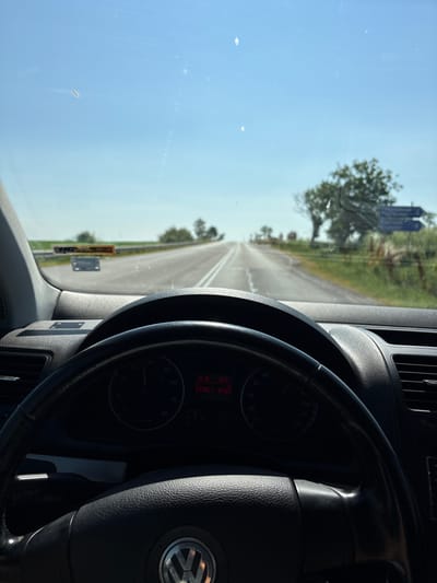 The image captures a first-person perspective from the driver's seat of a car, looking forward through the windshield. The road ahead is a two-lane asphalt road stretching into the distance, bordered by green fields and sparse trees under a clear blue sky. The car's dashboard is visible in the foreground, including the steering wheel with a Volkswagen logo and a speedometer. The time on the dashboard reads "10:18". A license plate or sticker is affixed to the lower portion of the windshield. The scene appears to be a daytime drive through a rural area. The location is identified as Zvegor, Bulgaria.