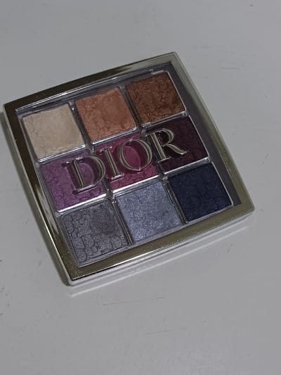 A square Christian Dior eyeshadow palette, enclosed in a silver-toned metallic compact, is positioned on a uniform, light gray or off-white surface. The palette contains nine individual eyeshadow pans arranged in a 3x3 grid. The top row features, from left to right, a light cream shade, a shimmery light bronze, and a shimmery dark copper. The middle row displays a shimmery purple, the raised silver "DIOR" logo centered on a vibrant fuchsia-pink shade, and a shimmery deep burgundy. The bottom row consists of a shimmery charcoal gray, a shimmery light silver-blue, and a shimmery dark navy blue. All eyeshadows exhibit a textured, pressed powder appearance with a shimmery finish. The underlying surface presents subtle, faint linear scuff marks. The scene depicts an indoor environment with even, bright illumination. No people, actions, or specific geographical identifiers for 서대문구, South Korea, are present within the frame.