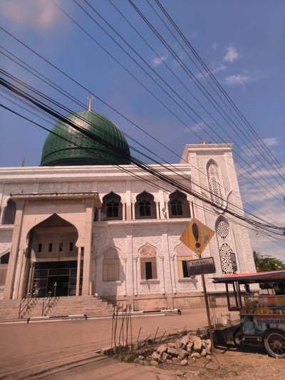 The image displays a large, white mosque featuring a prominent green dome crowned with a golden crescent moon finial, set against a vibrant blue sky with scattered clouds. Numerous black power lines cut across the upper part of the frame.

The mosque exhibits intricate architectural details, including ornate patterns carved into its white facade and a tall, slender minaret visible on the right. The main entrance is a grand arched structure with stairs and metal railings, appearing to be in an unfinished or renovated state with exposed concrete sections. Windows throughout the building also feature arched designs.

In the foreground, a dirt or gravel street runs alongside the mosque. On the right side, a yellow diamond-shaped sign with an exclamation mark is visible, mounted on a pole, with a black rectangular sign below it bearing the Indonesian word "KURANGI" (meaning "Reduce" or "Decrease"). Adjacent to the signs, a colorful, three-wheeled street vendor cart is partially visible, featuring text that appears to read "RUJAK SALAK PLU," suggesting it sells Rujak Salak (a fruit salad dish). Nearby, there is a pile of construction debris or bricks. Low, black and white striped barriers are positioned on the ground in front of the mosque steps; one barrier has the text "STOP SANDAL SEP," likely indicating a shoe-free zone before entering the mosque.

The scene is illuminated by bright daylight, suggesting a sunny day, likely late morning or early afternoon, in Keudelapang, Indonesia. There are no people visible, but the presence of the vendor cart and the construction materials hint at everyday activity around the mosque.