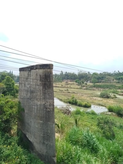 A slightly elevated perspective in Đại Ninh, Vietnam, captures a weathered, concrete structure positioned prominently in the foreground. The structure, possibly a section of a bridge or building support, displays vertical streaking indicative of water runoff and age. Wires run horizontally across the top portion of the frame, intersecting the concrete structure. Lush green vegetation surrounds the base of the structure and extends into the midground, where a body of water meanders through the landscape. In the background, a mix of grassy terrain and trees defines the horizon line under an overcast sky. No people are visible.