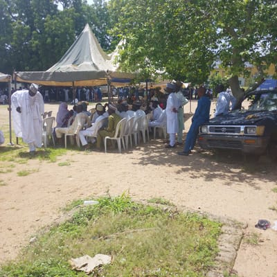 The image depicts an outdoor gathering under a large tent, likely in Gwoza, Nigeria. Several men are seated on white plastic chairs arranged in rows, while others stand or walk around. Many of the individuals are wearing traditional Nigerian attire, including long white robes and patterned caps. A black pickup truck is parked to the right of the scene, partially obscured by trees. The ground is sandy with patches of green grass and scattered vegetation. In the background, a building with yellow siding is visible through the trees. The overall atmosphere suggests a community event or a religious gathering.
