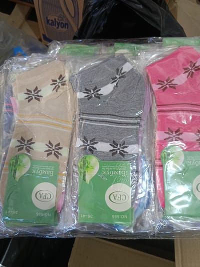 The image shows three pairs of socks packaged in plastic. The socks are in different colors: beige, gray, and pink, and each pair has a floral pattern. The packaging includes green labels with text that appears to be in Cyrillic and Latin alphabets, along with images of bamboo. The text includes "Bamoyk," "High-stretch new bamboo fiber," "Let your feet a touch of healthy," "CFA," "NO: 555," and "36-41." The socks are arranged side-by-side, and the packaging is transparent, allowing the socks to be clearly visible. In the background, out of focus, there are shelves or boxes, one of which has a label with the word "kalyon." This scene is likely from a retail environment in Berdiansk, Ukraine, given the Cyrillic text and the general context of packaged goods. There are no people or animals present in the image. The lighting is even, and the focus is on the socks and their packaging.