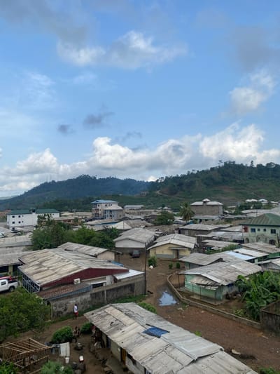 The image shows a residential area in Bonadikombo, Cameroon, with a large green hillside in the background. The view is from a rooftop, looking down on the rooftops of the houses. The sky is clear, with some clouds, and there is a small puddle in the dirt road in the foreground. Several people are walking on the street in the foreground. The houses are mostly single-story, with corrugated metal roofs. Some of the houses have gardens in the front yard, and others have fences around them. There are several trees in the background, providing some shade for the houses.