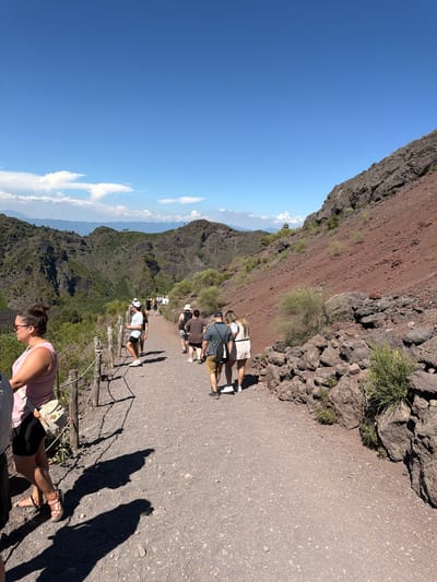 The image shows a group of tourists walking on a gravel path along the rim of a volcanic crater in Ercolano, Italy. The path is bordered by a wooden fence on the left and a steep, rocky slope on the right. The slope is covered in reddish-brown volcanic soil and scattered rocks. In the background, lush green mountains and valleys can be seen under a clear blue sky with scattered white clouds. The tourists are casually dressed for a warm day, with some wearing hats and sunglasses. They are walking in the same direction, seemingly on a guided tour. The overall scene depicts a popular tourist attraction with a natural volcanic landscape.