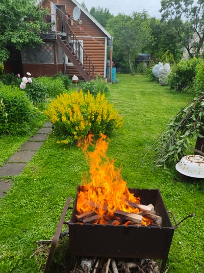 A bright orange fire burns in a dark metal brazier filled with wooden logs, positioned on a lush green lawn. To the left of the brazier, a stone pathway winds through vibrant green foliage and a bush covered in bright yellow flowers. In the background, a rustic wooden house with a satellite dish and an external staircase stands under an overcast sky. Various items are scattered around the yard, including blue barrels, large white bundles covered in plastic, and a weathered white basin with branches resting on it. The scene evokes a sense of a relaxed outdoor gathering or a backyard clean-up in a rural setting. The time of day appears to be daytime, with the cloudy sky suggesting potentially cooler weather. The location is identified as Автомобилист Снт, Russia. No text is visible in the image.