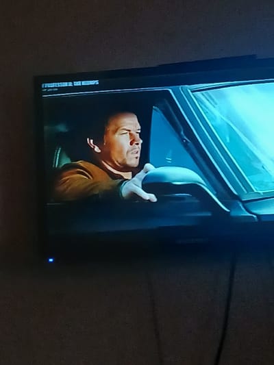 A flat-screen television, featuring a black bezel and a glowing blue power indicator light on its bottom-left, displays a video scene. The primary subject in the video is a man, visually resembling actor Mark Wahlberg, depicted in profile, seated in the driver's position of a vehicle. He wears a brown or olive-drab jacket, with his right hand resting on the steering wheel. His head is turned slightly to the right, and his facial expression appears serious or focused. The vehicle's interior, including parts of the dashboard and the front windshield, are visible, with diffused bright light appearing through the windshield. Partially legible text, possibly "FIGHT RESILIEN" or similar, appears in the top-left corner of the screen's content. The immediate environment surrounding the television is dark and indistinct, suggesting a dimly lit room or dark background. No visual information within this image establishes the physical location of the television set as being in the city of Otta, Nigeria.