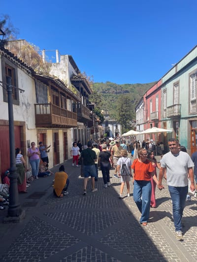 The image depicts a street scene in the city of Teror, Spain, on a sunny day. A diverse group of people are walking along a cobblestone street lined with colorful buildings. On the left, traditional Canarian architecture with wooden balconies is visible, while on the right, buildings are painted in vibrant hues of pink and turquoise. Several people are casually dressed, suggesting a leisurely atmosphere. Some are walking in groups, while others are alone. A street lamp stands on the left side of the image. In the background, a church with a clock tower and mountainous terrain can be seen under a clear blue sky.