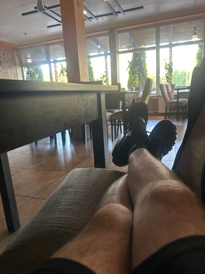A first-person perspective shot captures a person relaxing indoors, with their legs crossed and resting on a textured chair. The person is wearing black athletic shoes and shorts, with visible leg hair. The setting appears to be a cafe or restaurant, suggested by the presence of tables and chairs visible through a large window. Outside the window, lush greenery and trees are visible, bathed in bright sunlight. The indoor environment features a large, dark wooden table in the foreground and light-colored tiled flooring. The decor includes a tall, light-brown pillar and subtle wall patterns. The overall impression is one of a tranquil moment in a well-lit, pleasant establishment, possibly in a suburban or rural setting, given the prominent natural elements outside. The location is identified as Zapolyev, Russia.