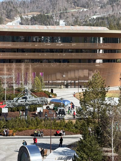 The image shows the "Горный воздух" leisure center in Озёрное, Russia, on a sunny day. A modern building with horizontal wooden slats on its façade stands tall. A chairlift station is visible in the foreground, with chairs suspended above a pathway. People are sitting on benches along the pathway, and a few individuals are walking. Trees and landscaping add greenery to the scene. In the background, a snow-covered mountain or hill is visible, likely part of the skiing area.
